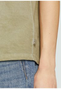 T-shirt en coton vert olive avec un ourlet incurvé, une texture subtile et une petite étiquette au bas de la couture ; assortie à un jean bleu délavé.