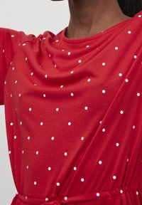 Punainen puuvillapaita, jossa on valkoisia pilkkuja, löysällä istuvuudella ja kerätyllä vyötäröllä säätötyylin mahdollistamiseksi.