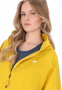 Sárga vízálló dzseki cipzárral, állítható kapucnival és fehér esernyő logóval. Sötétkék-fehér csíkos ing fölött viselve. Simább textúra.