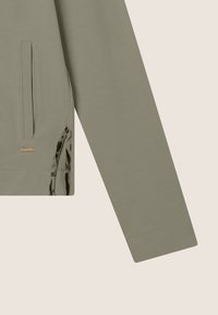 Sweatshirt vert olive à manches longues, avec poches latérales et un accent texturé sur l'ourlet inférieur présentant un motif. Détail du logo doré inclus.