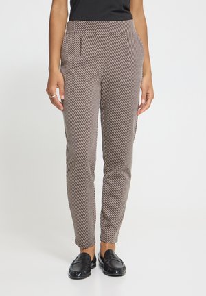 Personne portant un pantalon taille haute, ajusté, à rayures diagonales marron et blanches, avec des mocassins noirs et un haut noir, les mains détendues sur les côtés.