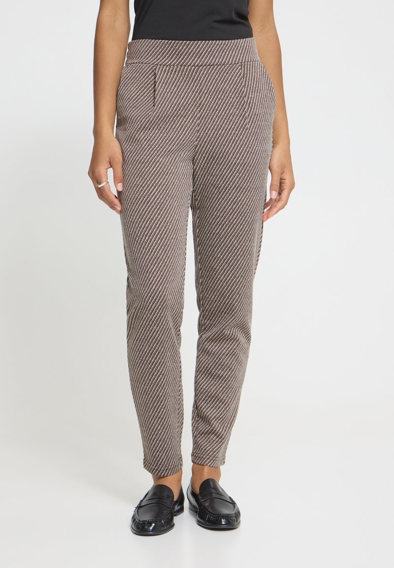 Personne portant un pantalon taille haute, ajusté, à rayures diagonales marron et blanches, avec des mocassins noirs et un haut noir, les mains détendues sur les côtés.