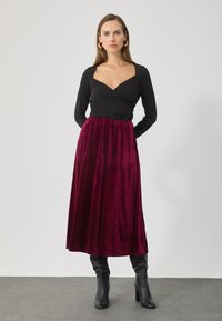 Top negru cu mânecă lungă și decolteu în formă de V, asortat cu o fustă burgundy din catifea, lungime ankle. Modelul poartă cizme negre până la genunchi.