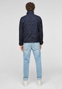 Mörkblå lättviktsjacka med ståkrage, detalj i bakre sömmen och knäppta muddarna, kombinerad med ljusblå jeans och vita sneakers.