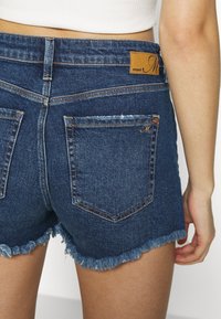 Vista trasera de una persona con pantalones cortos de mezclilla desgastados de color azul oscuro y una blusa corta blanca, con la mano relajada a un lado.