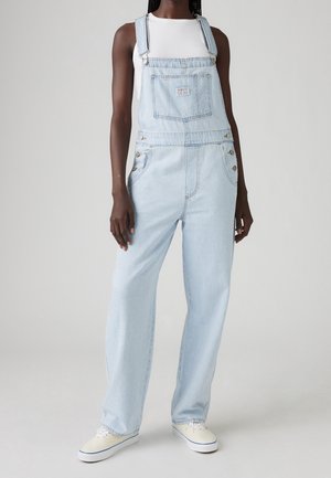 Osoba o ciemnej karnacji ubrana w jasnoniebieskie denimowe ogrodniczki, biały bezrękawnik i białe trampki, stojąca na tle jednolitego jasnego tła.