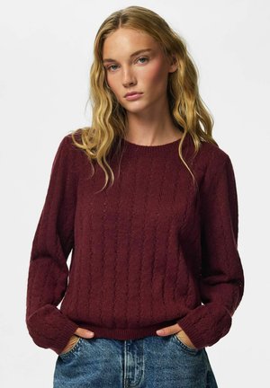 Femme aux longs cheveux blonds ondulés, portant un pull torsadé couleur bordeaux et un jean bleu, debout les mains dans les poches devant un fond uni.