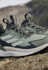 Zeleno-černé outdoorové boty s texturovaným povrchem, s třemi bílými pruhy, štítkem GORE-TEX a úchyty.