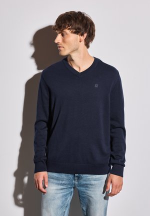 Jersey de cuello en V en azul marino, hecho de un material de punto suave, con mangas largas, puños y dobladillo acanalados, y un pequeño logo en el pecho.