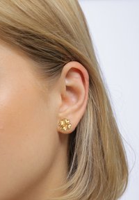 Goldene Ohrstecker in Blütenform mit strukturiertem Design und sanften Kurven, vor einem hellen Hintergrund.