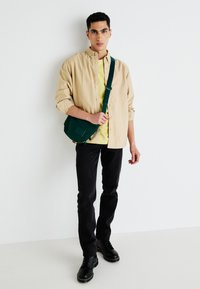 Beige langemouwen shirt, groene schoudertas, gele t-shirt, zwarte jeans en zwarte veterschoenen, staand tegen een witte achtergrond.