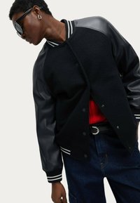 Chaqueta varsity negra con cuerpo de forro polar y mangas de cuero, que presenta puños acanalados a rayas y una capa de punto roja debajo.