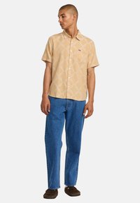 Short-sleeve beige shirt featuring a subtle patterned design, paired with blue denim jeans and brown footwear.