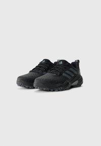 Baskets de sport noires Adidas avec des semelles texturées et trois rayures grises sur le côté, présentées sur un fond blanc uni.