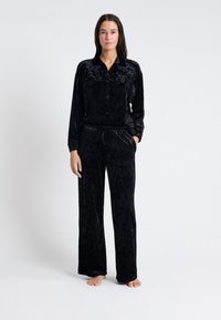 Conjunto de loungewear de terciopelo negro que incluye una blusa de manga larga con botones y pantalones a juego con cordón. Textura suave y corte holgado.
