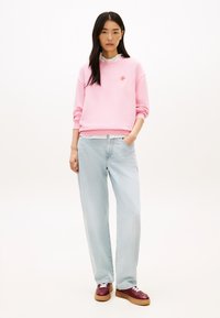 Ροζ φούτερ με ριμπ μανσέτες και στρογγυλή λαιμόκοψη, με μικρό κόκκινο λογότυπο. Ανοιχτό μπλε τζιν παντελόνι με φαρδιά μπάσκα και μπορντό αθλητικά παπούτσια με λευκά κορδόνια.