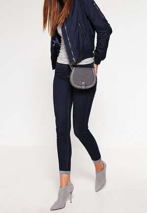 Femme portant une veste bomber bleu marine, un haut gris, un jean skinny foncé, des bottines grises à talons hauts et un petit sac gris porté en bandoulière.
