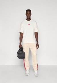 Hellbeiges Sportoutfit mit roten Akzenten, bestehend aus einem kurzärmeligen Oberteil und Hosen mit Seitenstreifen. Inklusive grauer Sporttasche.