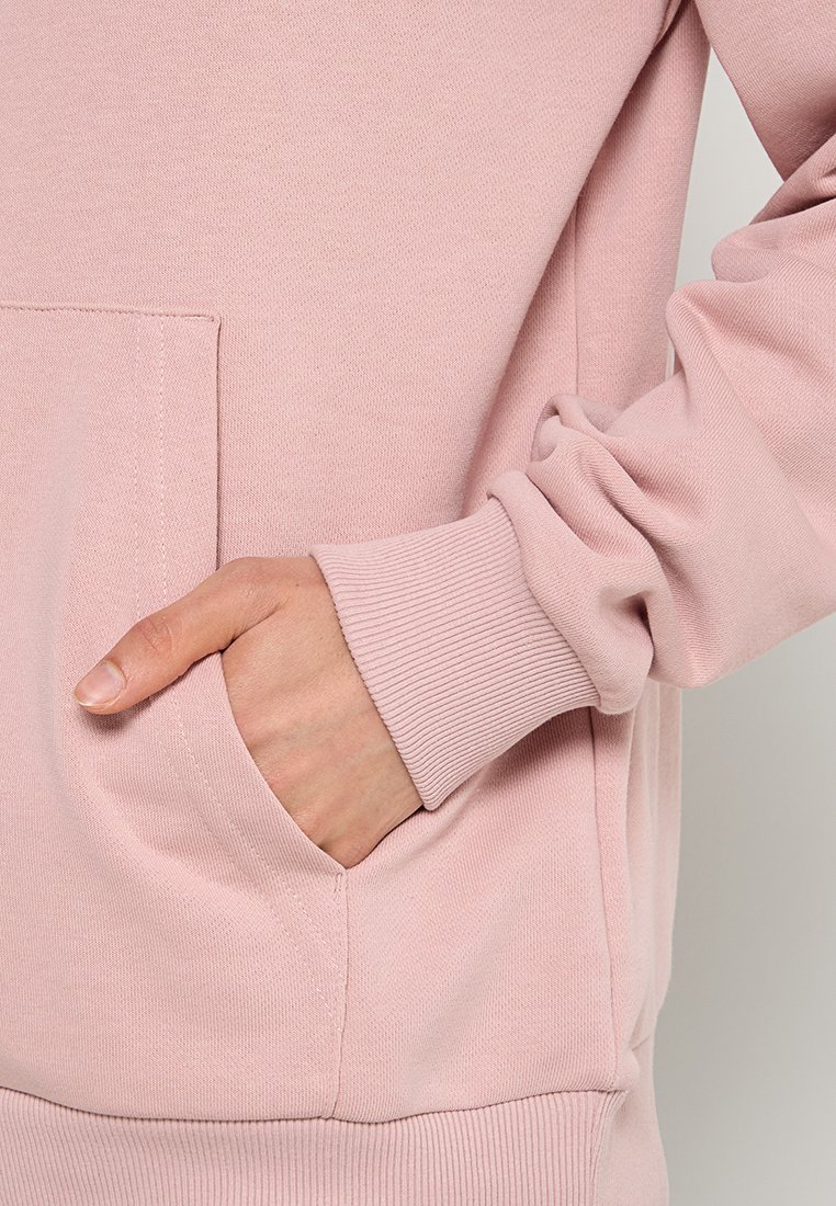 Lichtroze sweatshirt met ribbelmanchetten en tailleband. Voorzien van een kangaroozak en zachte, gestructureerde stof. Close-up van hand in de zak.