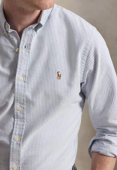 Homme portant une chemise boutonnée à rayures bleu clair et blanches, avec les manches retroussées et un petit logo brodé de cheval et cavalier sur la poitrine.