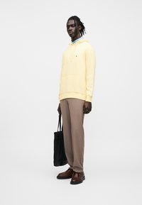 Κίτρινο φούτερ με τσέπη τύπου καγκουρό, συνδυασμένο με καφέ παντελόνι και καφέ παπούτσια. Κρατάει μια μαύρη τσάντα tote. Ομαλή υφή υφάσματος.