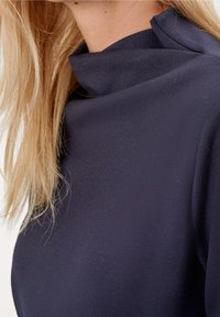 Top blu navy con collo alto realizzato in tessuto liscio. Presenta una silhouette pulita con accenti minimi e una finitura testurizzata discreta.