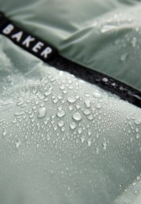 Material textil verde deschis, rezistent la apă, cu o textură netedă, având picături de apă. Bandă neagră cu logo-ul "BAKER" vizibil.