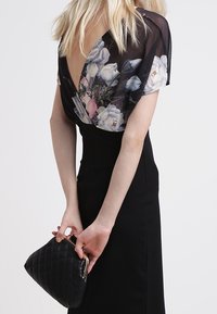 Černé šaty s květinovým vrchním dílem ve stylu zavazování s hlubokým V-zadním výstřihem. Modelka drží černou prošívanou kabelku na řetízku s kovovým zapínáním.