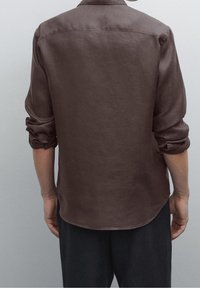 Braunes Langarmhemd aus strukturiertem Stoff mit Rückenpasse, hochgekrempelten Ärmeln und abgerundetem Saum, getragen mit dunkler Hose.