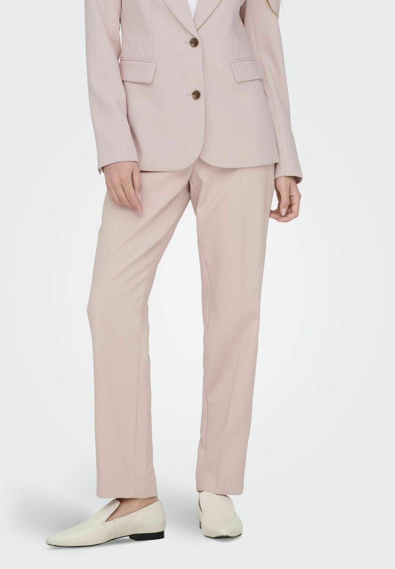 Traje rosa claro con blazer ajustado que presenta dos botones y bolsillos laterales, combinado con pantalones rectos a juego y zapatos de corte ligero.
