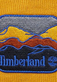Gult tyg med ett blåbroderat märke som har ett bergslandskap i grått, orange och gult, med ordet "Timberland" nedanför.