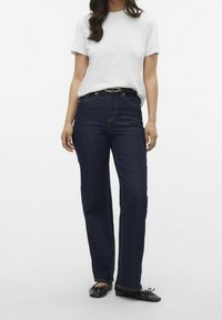 Jeans blu scuro con una vestibilità a gamba dritta, vita alta e impunture gialle, abbinati a una semplice t-shirt bianca a maniche corte e ballerine nere.