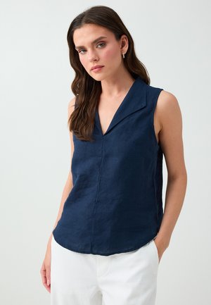 Junge Frau mit langen dunklen Haaren trägt eine ärmellose marineblaue Bluse und weiße Hose, steht mit einer Hand in der Tasche.