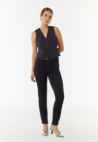 Marineblaues, schmal gestreiftes Weste mit V-Ausschnitt und fünf Knöpfen, kombiniert mit dunklen Jeans mit gelben Nähten und silbernen High Heels.