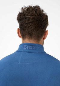 Polo blu con superficie texturizzata, caratterizzato da un logo "Brax" in rilievo sul colletto. Il design include un colletto strutturato e uno stile minimalista.
