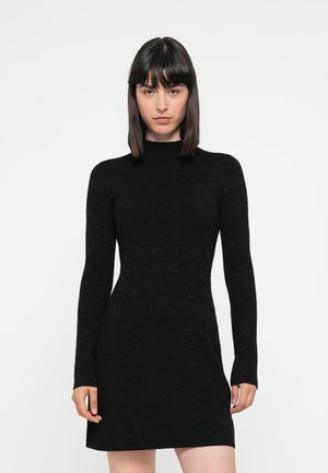 Woman with straight dark hair wearing a fitted black long-sleeve dress with subtle textured pattern, standing against a plain white background.