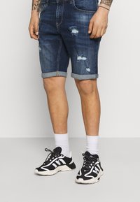 Denim shorts in dark wash with distressed details, rolled hem, paired with black and white sneakers and white crew socks.
