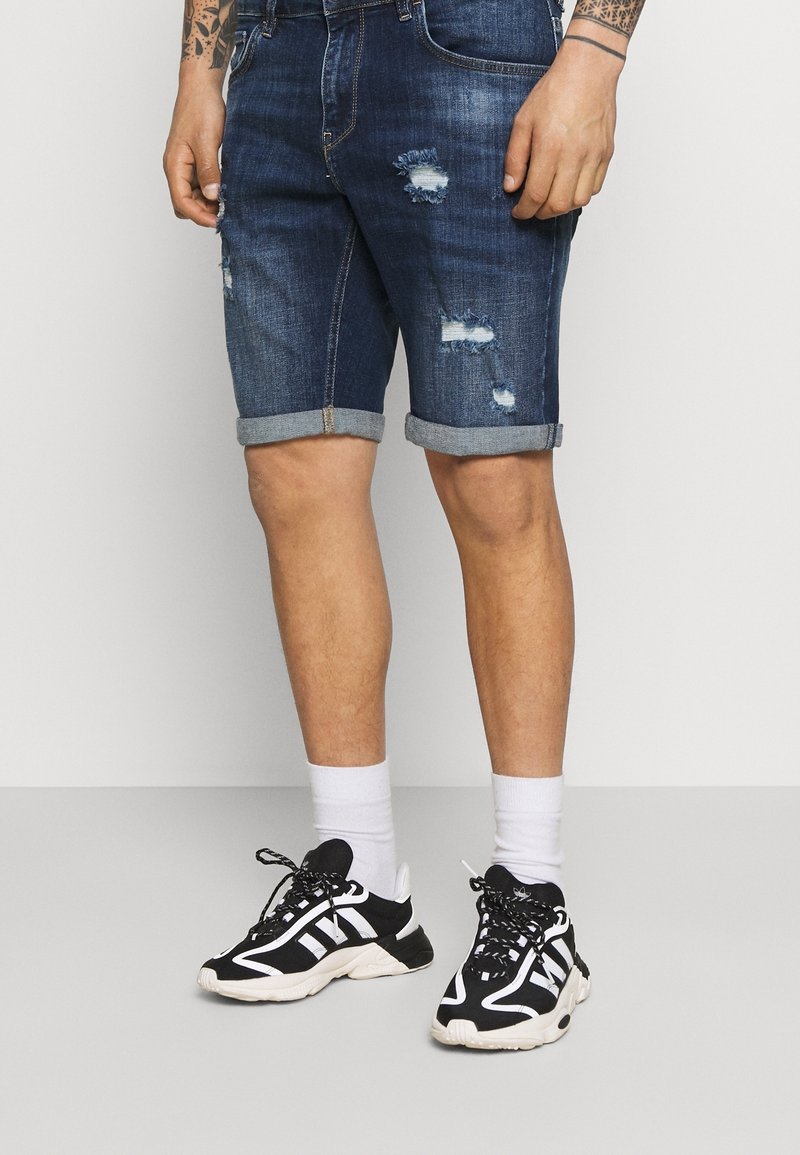 Denim shorts in dark wash with distressed details, rolled hem, paired with black and white sneakers and white crew socks.