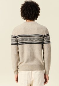 Suéter de punto beige con cuello redondo, que presenta un patrón horizontal oscuro en el pecho. Puños y dobladillo acanalados, textura suave.