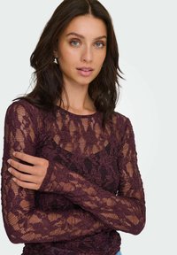 Top de encaje burdeos de manga larga con patrones florales, textura semitransparente y escote redondo. Las mangas llegan hasta las muñecas.