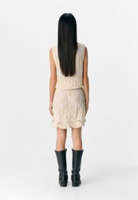 Beiges, ärmelloses Kleid mit aufwändiger Stickerei und gewelltem Saum; kombiniert mit knielangen schwarzen Lederstiefeln. Rückansicht angezeigt.