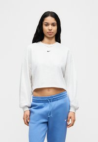 Ανοιχτό γκρι κοντοκουρεμένο φούτερ με μακριά μανίκια και στρογγυλή λαιμόκοψη, με μικρό μαύρο λογότυπο Nike. Συνδυάζεται με ανοιχτό μπλε παντελόνι φόρμας.