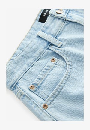 Vaqueros de denim azul claro con un primer plano de la cintura, que presenta un botón, remaches y un pequeño bolsillo para billetes. Textura sutil visible.