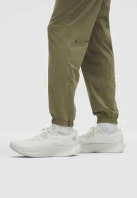 Weiße Sportschuhe mit strukturierter Oberfläche, kombiniert mit hochgerollten olivgrünen Jogginghosen, die Knöchelsichtreissverschlüsse und elastische Bündchen haben.