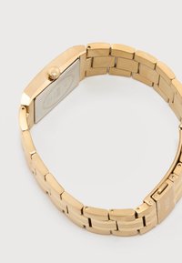 Ρολόι χειρός από μέταλλο σε χρυσό τόνο με συνδεδεμένο μπρασελέ και ορθογώνια θήκη, εμφανισμένο από πίσω σε λευκό φόντο.