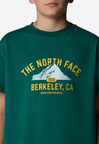 Green cotton t-shirt featuring a mountain graphic and text: "THE NORTH FACE," "BERKELEY, CA," and "NEVER STOP EXPLORING" in yellow.