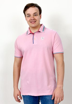 Jeune homme souriant, portant un polo rose clair avec col et bords de manches rayés, debout devant un fond blanc uni.