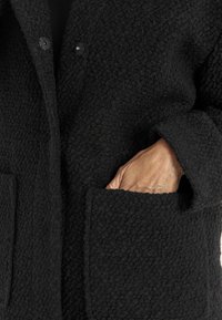 Hand teilweise in der Tasche eines strukturierten schwarzen Mantels mit sichtbaren Knöpfen und einem lockigen Gewebemuster.