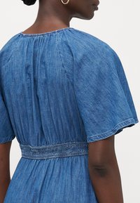 Vestido de mezclilla con mangas cortas, cintura ajustada y detalles fruncidos. Presenta un escote fruncido y una tela suave y texturizada en un tono azul medio.