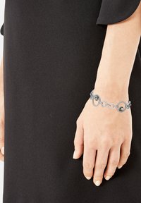 Pulsera de cadena de plata con eslabones circulares y dos piedras oscuras, llevada en una mano sobre un fondo negro sólido.